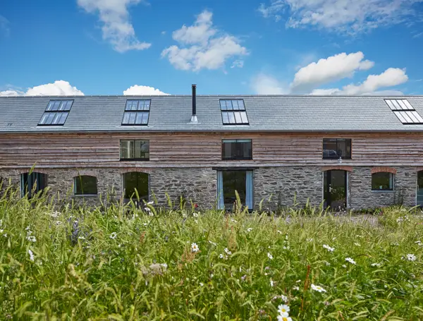 Converted stone barn with wooden cladding, featuring multiple pitched rooflights on the sloped roof, set against a backdrop of a clear blue sky.